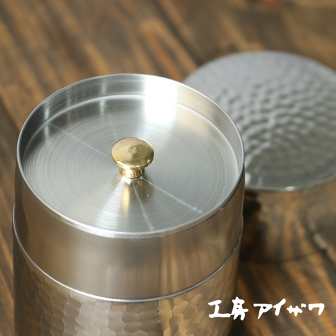 銀色不鏽鋼錘目紋茶葉罐，配有金色圓形提手，罐身有明顯的錘擊紋理，另一同款茶葉罐蓋子在背景中模糊顯示。