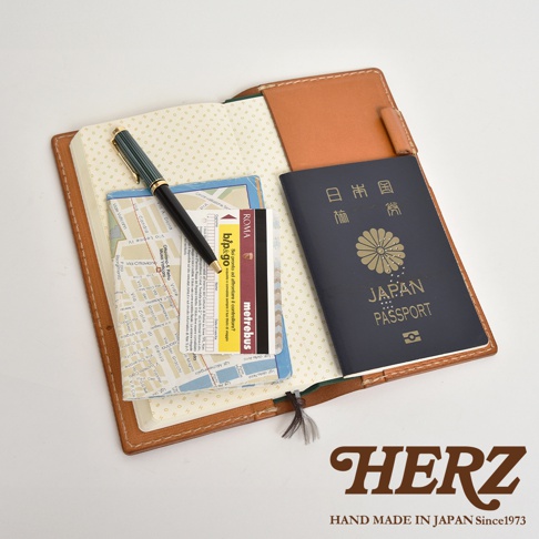 職人手作皮革旅行記事本書套，展示一個打開的淺棕色皮革封套，內含一本點陣筆記本、一張地圖、數張卡片及一本深藍色日本護照。