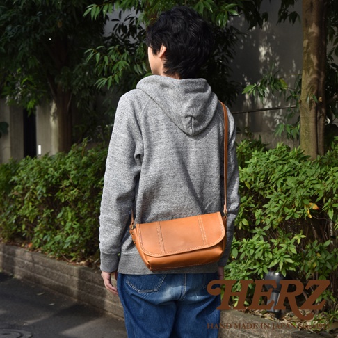 一個身穿灰色帽 T 和藍色牛仔褲的人，斜挎著一個棕褐色皮革復古郵差包。包包設計簡潔，有明顯的縫線細節，風格休閒。