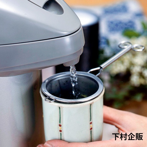 不鏽鋼深型濾茶網，網孔細緻，附有長柄，正置於有綠色與紅色裝飾的杯口，上方有熱水從銀色電熱水壺中流出，濾網邊緣有兩個掛環。