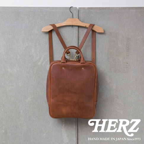 一個棕色的皮革後背包，掛在木製衣架上，背景是灰色牆壁，右下角有HERZ品牌標誌。