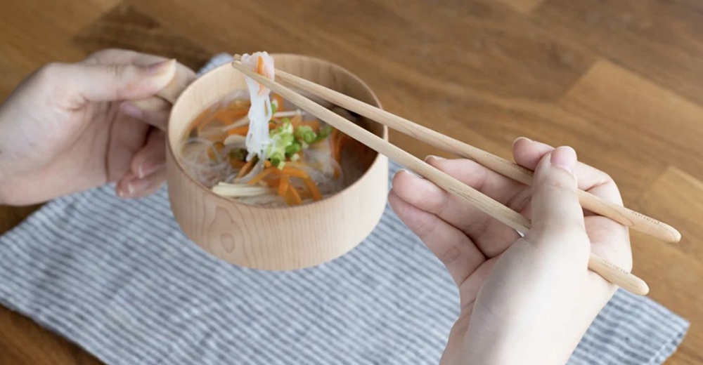 一雙淺木色楓木圓筷，長度約 21 公分，夾起碗中湯麵，碗旁有條紋餐巾。