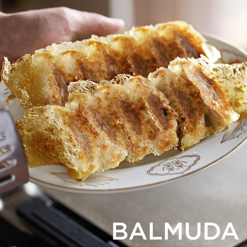 盤中盛有金黃酥脆的煎餃，部分煎餃底部帶有網狀焦脆層。盤子邊緣有金色裝飾圖案，背景可見一隻手和產品品牌名稱「BALMUDA」。
