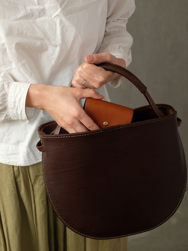 職人手作咖啡色皮革迷你短夾，以圓潤的袋身設計，搭配咖啡色皮革手柄，整體呈現自然質樸的風格，細緻的縫線工藝增添質感。