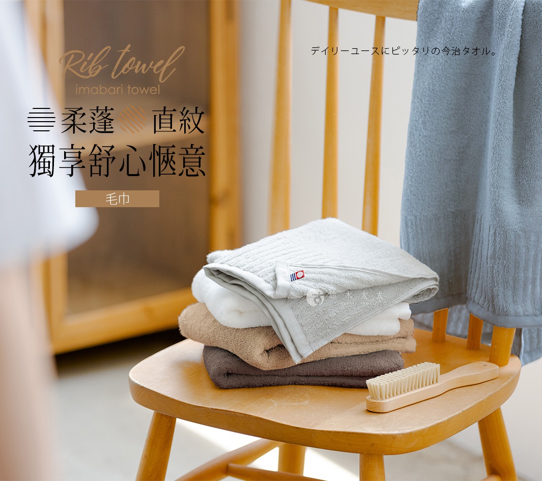疊放的今治毛巾，最上方為淺灰色，帶有直紋與品牌標誌，下方依序為白色、淺棕色和深棕色毛巾，旁邊擺放一把木柄小刷子，毛巾堆疊在木椅上。