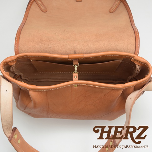 職人手作皮革郵差包，採用天然棕色皮革，包身線條簡潔，內部有置物夾層，搭配金屬扣環與皮革背帶。