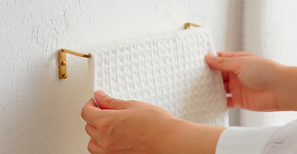 黃銅單桿毛巾架，固定在白色紋理牆面上，一塊白色格紋毛巾搭在上面，一隻手正在整理毛巾。