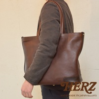 一個身穿棕色外套的人側身背著一個深棕色皮革托特包，包包上有拉鍊和皮革提把，包包底部有強化設計。