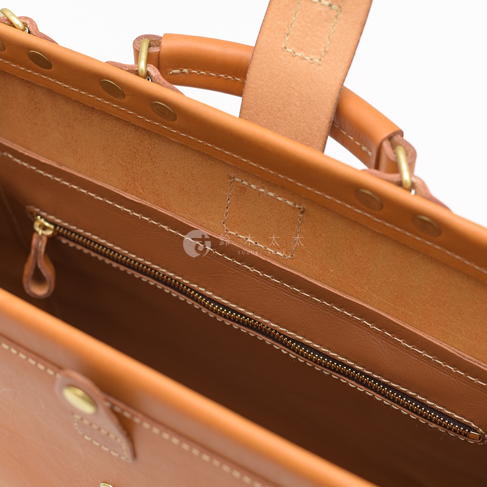 職人手作皮革軟式杜勒斯包，呈現自然棕色皮革質感，內部細節包含拉鍊、皮革拉環、車縫線及金屬卯釘，整體設計簡約實用。