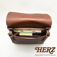 職人手作皮革經典郵差包，深棕色，內部裝有書籍、平板電腦和筆記本，展現皮革材質的質感與實用性。