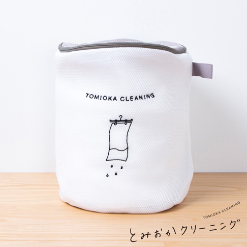 一個白色網狀材質的圓筒形洗衣袋，頂部有灰色拉鍊和一個灰色提把。袋身印有黑色「TOMIOKA CLEANING」字樣，下方是一個手繪圖案，描繪一塊毛巾掛在衣架上滴水。