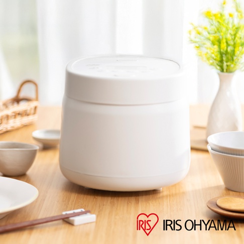 一個白色的電子鍋，側面印有 IRIS OHYAMA 的標誌，擺放在木質桌面上，背景是白色窗簾和一束黃色花飾。