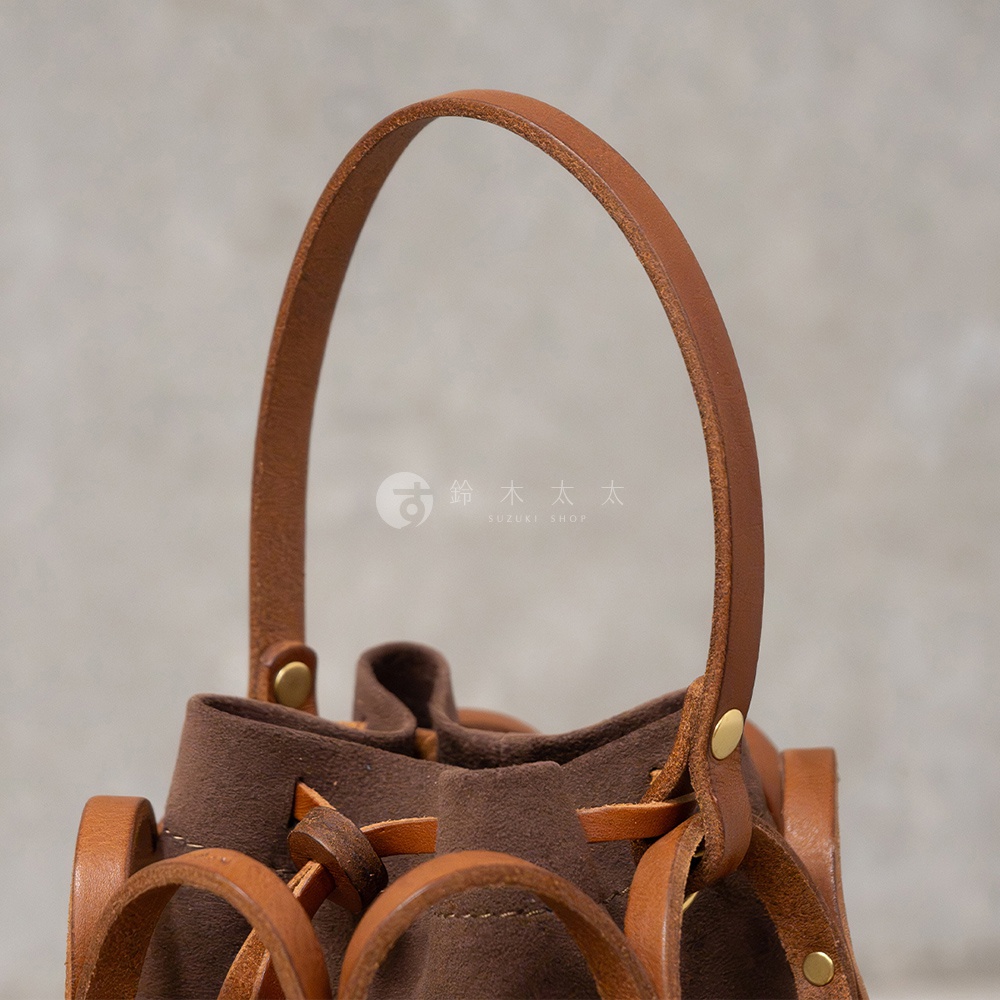 這是一款HERZ品牌日本職人手作皮革Organ編織水桶包，採用棕色絨面革和皮革材質，特寫鏡頭展示了包包的提把和抽繩設計。