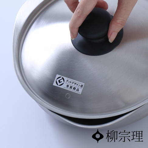 柳宗理品牌的不鏽鋼霧面單手鍋，上方覆蓋著霧面不鏽鋼鍋蓋，鍋蓋中央有黑色圓形把手，手部正握持把手。鍋蓋表面貼有標註著日文產品獲獎資訊的白色貼紙，右下角則印有柳宗理品牌名稱與標誌。