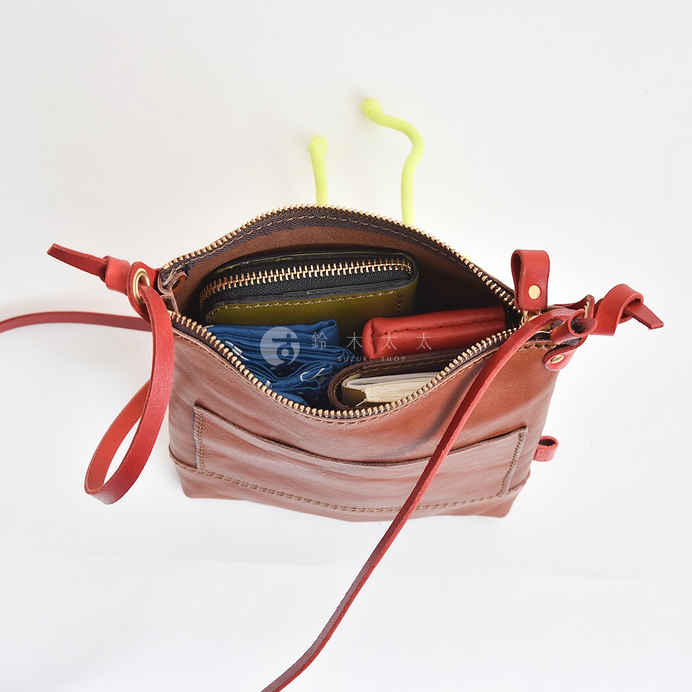 職人手作皮革直式隨身單肩包，棕色皮革材質，包內裝有綠色皮夾、紅色零錢包、藍色摺疊傘及筆記本。