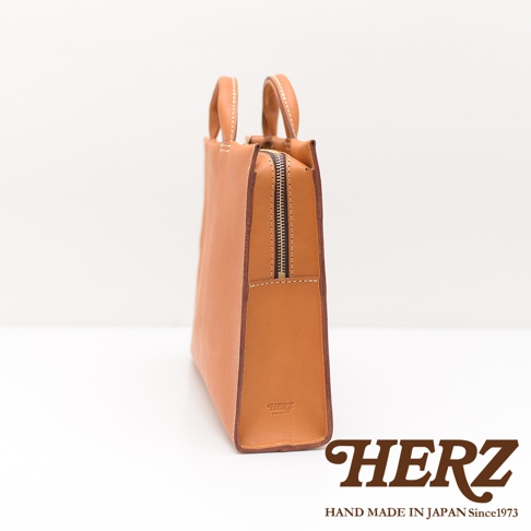 HERZ 品牌皮革公事包，採用駝色皮革製作，拉鍊設計，包包右側印有 HERZ 品牌標誌與 HAND MADE IN JAPAN Since1973 字樣