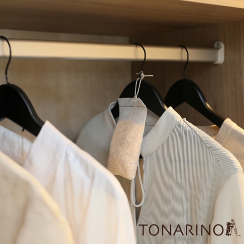 一個裝有淺褐色顆粒的透氣白色布包，懸掛在一組衣櫃中的黑色衣架上，旁邊是兩件淺色衣物。