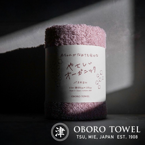 捲起的一條淡紫色有機棉浴巾，標籤上印有日文和英文說明，包括「OBORO TOWEL」品牌字樣，尺寸約 60 x 120 公分，100% 純棉，日本製造。