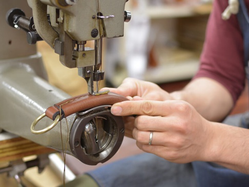 職人使用工業縫紉機，以手工縫製棕色皮革製品。皮革帶上裝有金色環扣，縫線清晰可見，展現精湛工藝。