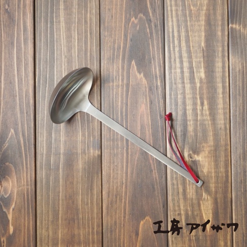 一把銀色不鏽鋼湯勺，勺柄末端繫有紅色編織吊繩，平放在深棕色木紋背景上。