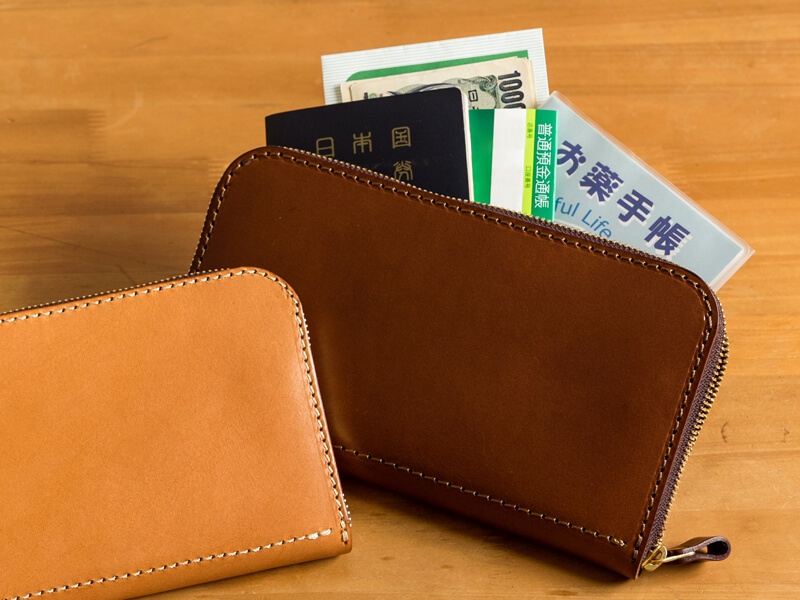 兩個職人手作皮革長夾，一個為淺棕色，另一個為深棕色，皆有拉鍊開合與手工縫線。深棕色長夾內裝有護照、日圓鈔票與藥手冊。