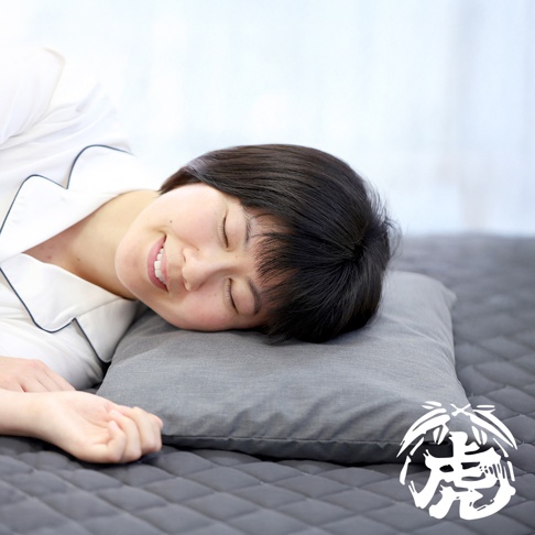 一位女性側臥在灰色枕頭上，枕頭平放在菱格紋床墊上。女性身穿白色睡衣，頭部輕靠在枕頭上，閉眼並露出微笑。