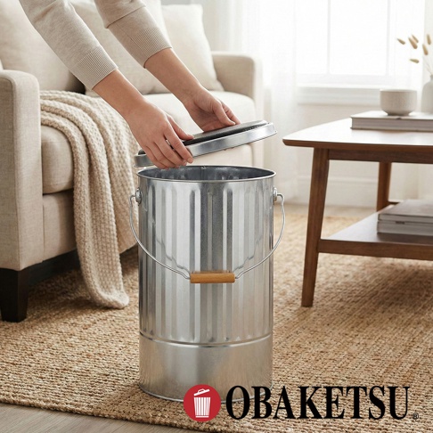 OBAKETSU 品牌的鍍鋅鋼手工儲物桶，桶身有立體壓紋，附木質提把，一位女士正將蓋子蓋上。