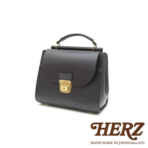 日本HERZ品牌黑色皮革手提斜背包，包身呈現穩重的瓦風造型，配有金色鎖扣與提把，下方有HERZ及HAND MADE IN JAPAN Since1973字樣。