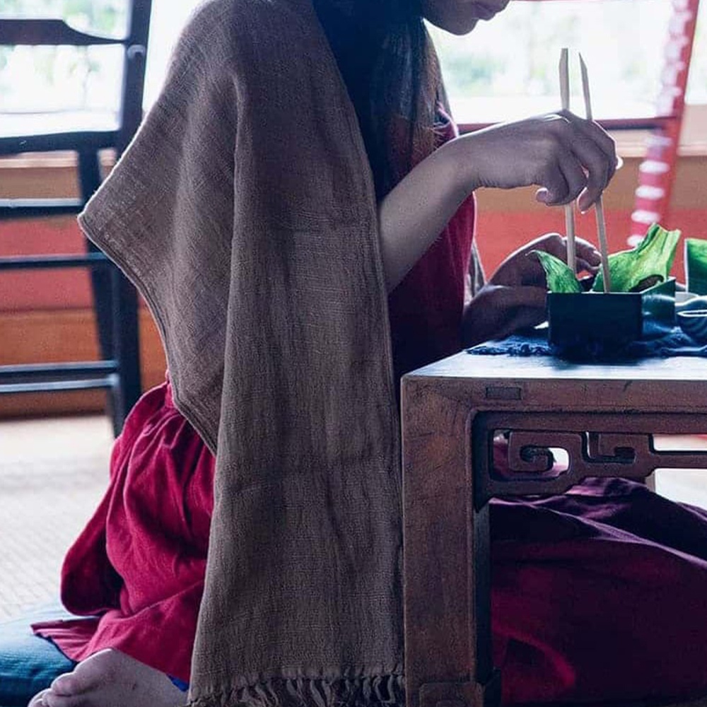 一位女士身穿紅色洋裝，披著一條棕色今治匹馬棉經典圍巾，圍巾帶有流蘇。她坐在木桌旁，用筷子夾取綠色植物。