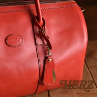 紅色皮革製成的手提包，帶有品牌標誌，以及一個皮革綁帶和金屬鞋拔吊飾，吊飾上飾有金屬扣和鉚釘。