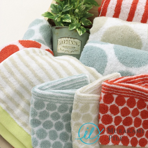 一盆綠植旁堆疊著色彩繽紛的毛巾，有條紋、圓點和純色的款式，部分毛巾上印有MURAKAMI PILE的品牌標誌。