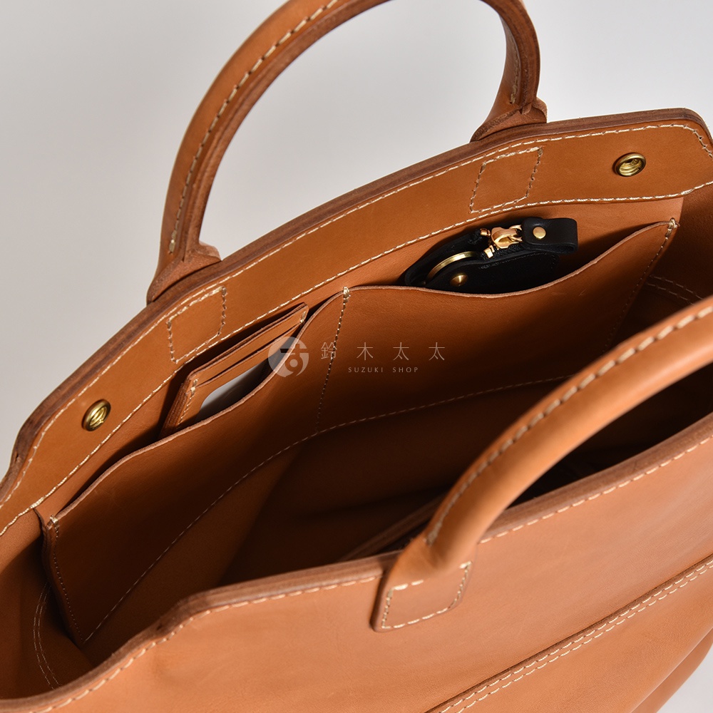 職人手作皮革大容量兩用托特包，採用光滑的駝色皮革製成，具有精緻的縫線細節。包包內部有兩個開放式隔層和一個帶鑰匙圈的拉鍊小袋，展現其大容量和實用性。
