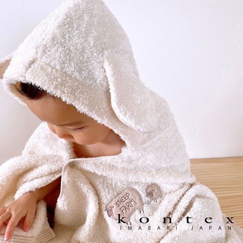 嬰兒穿著一件米白色、觸感柔軟的連帽浴巾，浴巾設計成可愛的動物造型，有耳朵裝飾。浴巾上繡有「SHEEP FARM」字樣和綿羊圖案。