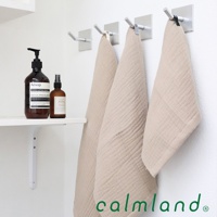 三條淺米色無撚紗質夾心長毛巾，質地柔軟，懸掛於白色牆面上的銀色掛鉤。左側架子上擺放著兩瓶棕色瓶身的清潔用品。