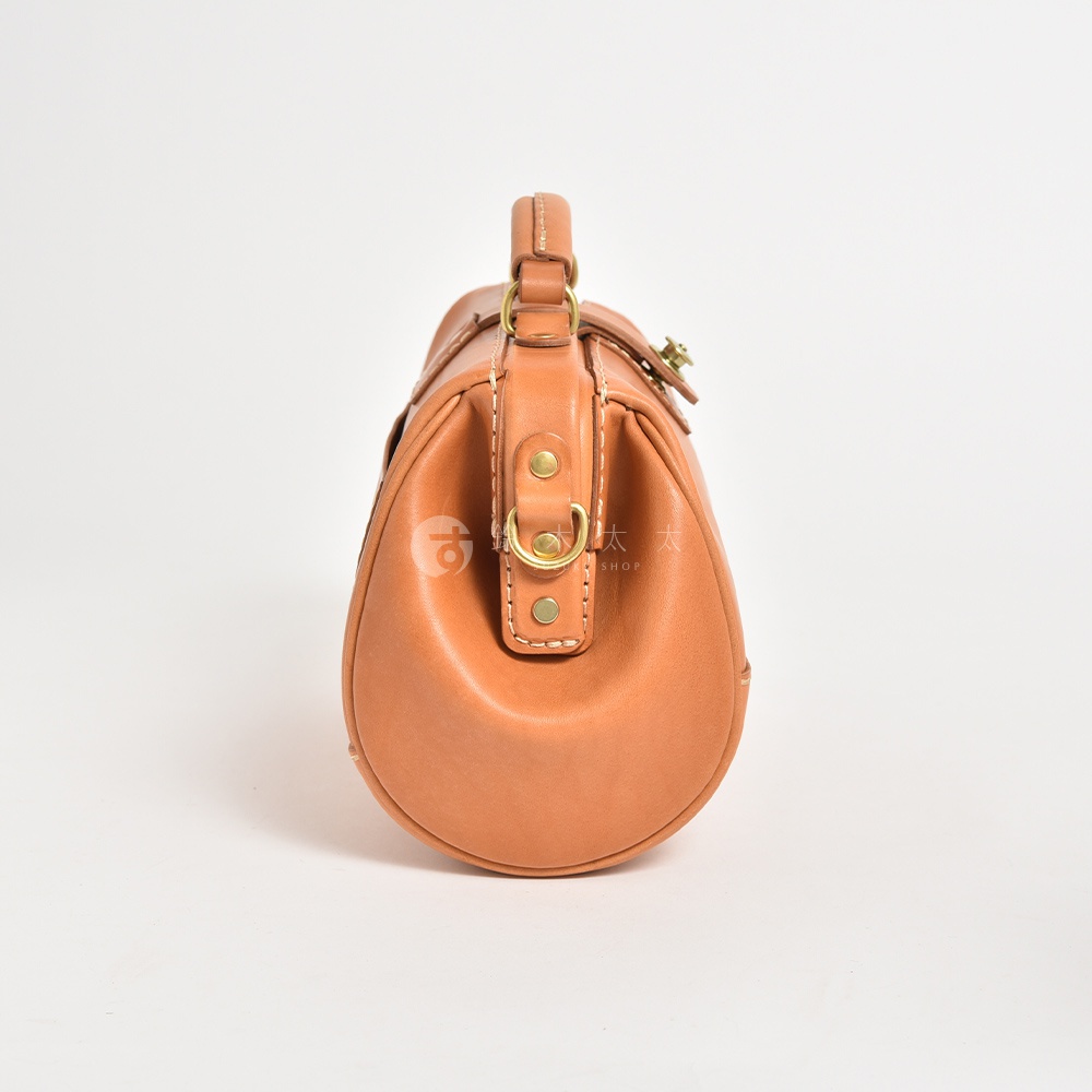焦糖色皮革製作的職人手作口金醫生包，配有黃銅色五金配件和提把，側邊展示包包圓潤的輪廓與車縫細節。