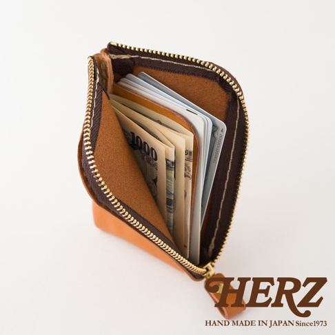 日本HERZ職人手作皮革迷你錢包，採用天然淺棕色皮革製成，錢包呈現半開口狀態，內部設有夾層可收納日圓紙鈔與卡片，配有金色拉鍊，整體設計簡約實用，右下方印有深褐色品牌名稱與產地字樣。