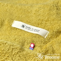 細節特寫：一條黃綠色、觸感柔軟的今治毛巾被，上面縫有品牌標籤，標籤上印有「made in Japan」和「La vie avec Broome」字樣，以及一個小巧的紅白色圓點和藍色條紋的裝飾標識。