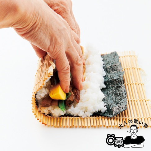 雙手正在竹簾上製作壽司捲，簾上鋪有海苔、白米飯和多種配料，包括黃色方塊狀的食物。
