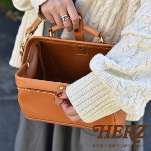 職人手作皮革口金醫生單肩包，呈現溫暖的焦糖棕色，採用厚實皮革製成，細緻縫線和金色口金細節，搭配象牙白麻花針織衫。