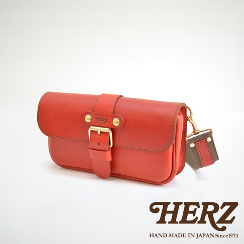 紅色皮革製成的單扣斜背包，包身帶有皮帶扣和品牌標誌，附有帆布與皮革拼接的肩帶。