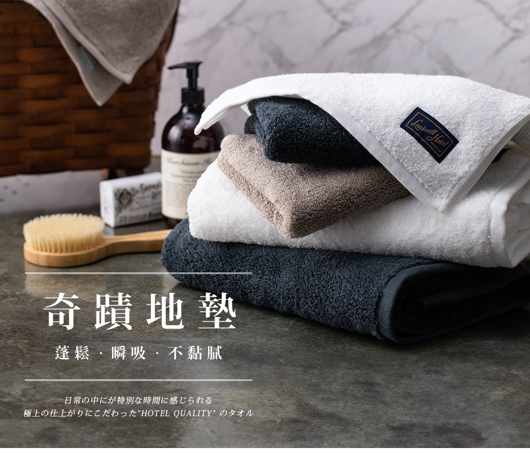 堆疊的泉州匹馬棉浴巾，包括深藍色、淺棕色和白色。白色浴巾上帶有藍色標籤，標有「Landwell Hotel」字樣。背景中可見洗浴用品和木柄刷。