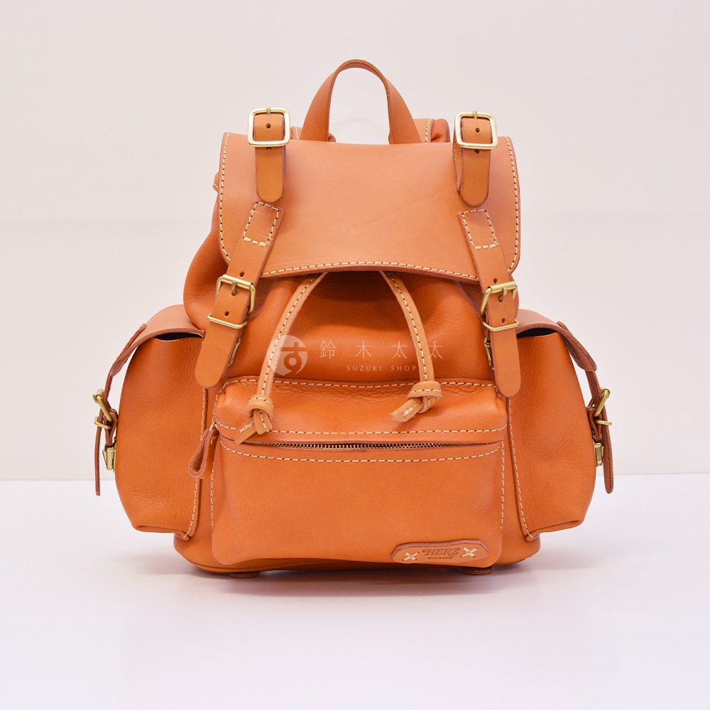 職人手作皮革復古風旅行後背包 R-65，採用天然皮革製成，呈現溫潤的橘棕色調，搭配金色金屬扣環和抽繩設計，側面有兩個小口袋，正面有一個拉鍊口袋，整體風格簡約而有質感。