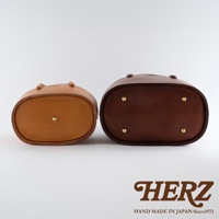 職人手作皮革束口手提包，左為淺棕色，右為深棕色，兩包均為橢圓形底部，配有四個黃銅腳釘，底部中央亦有一個黃銅釘。