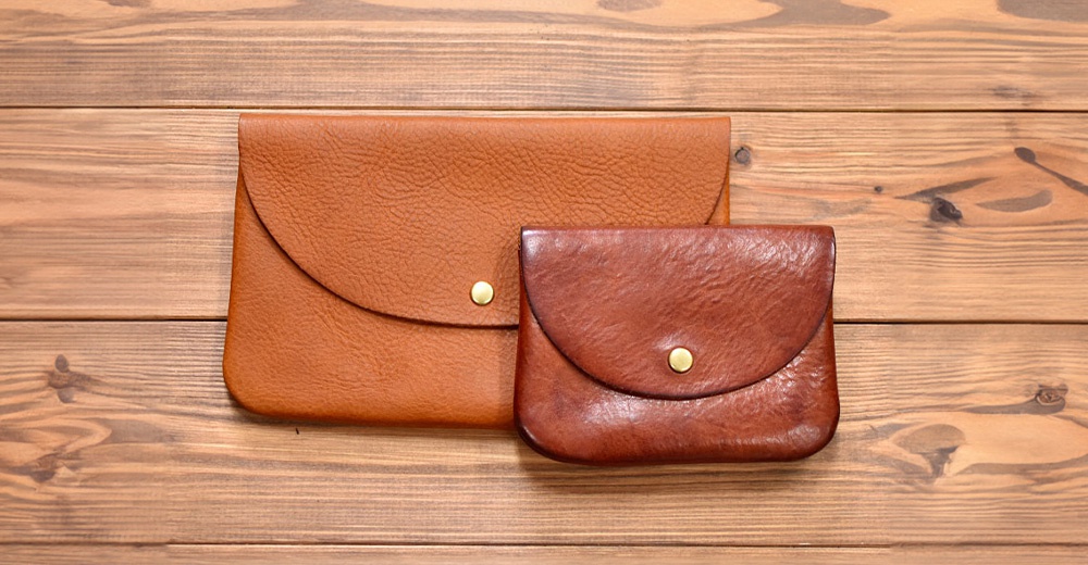 兩款職人手作皮革小物收納袋，左為淺棕色，右為深棕色，皆為鈕扣式設計，材質為皮革，置於木紋背景上。