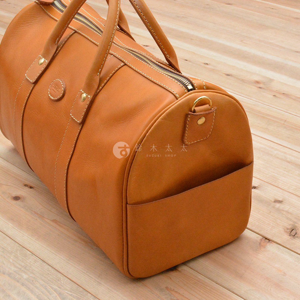 棕褐色皮革製成的圓筒形波士頓包，配有拉鍊開合、兩條提把，側面有一個開放式口袋，整體風格簡潔經典。