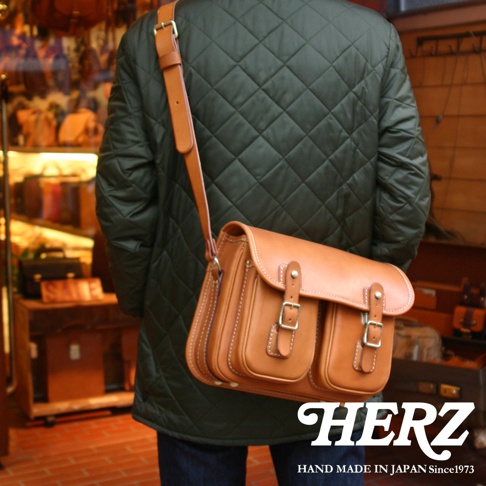 一個穿著綠色菱格紋外套的人，身後揹著一個褐色的經典雙扣郵差包，包包上有HERZ的品牌字樣，並寫著HAND MADE IN JAPAN Since1973。