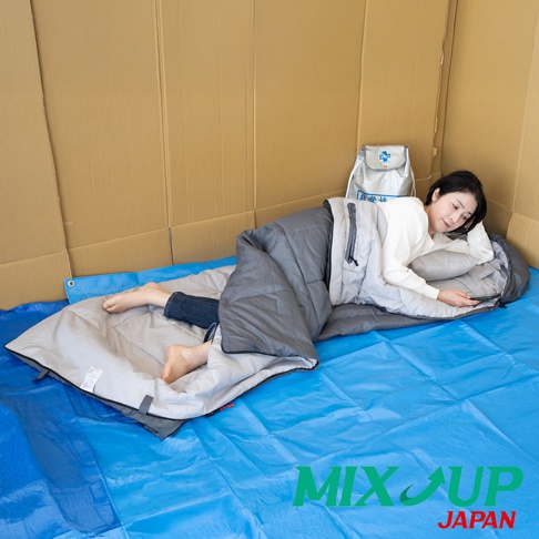 一個女人躺在睡袋裡，臉上帶著微笑，手機放在右手中，身旁有一個白色的背包，背景是紙板牆壁，地板上鋪著藍色的防水布，上面有MIX UP JAPAN的標誌。