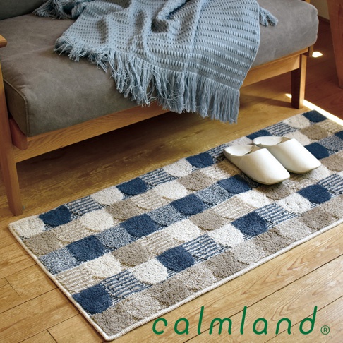 長方形地墊，採用藍色、米白色及大地色系棋格紋設計，材質為織物，上方擺放一對白色室內拖鞋，背景為木質地板及灰色沙發，沙發上搭有藍色流蘇毯子。