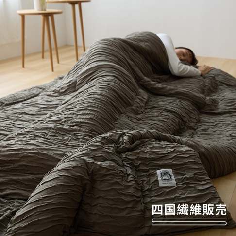 一位女子正躺在鋪在地板上的床鋪休息，身上蓋著一件深褐色的民族風褶皺紋理單人被，被子右下角縫有四國纖維的標籤，整體氛圍呈現出舒適與簡約的居家風格。