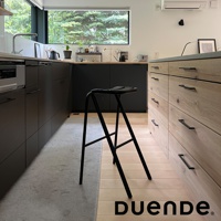 黑色金屬吧檯椅，椅面為一體成形設計，椅腳為細長金屬支架，整體造型簡約，擺放在木質地板與深色廚具之間。